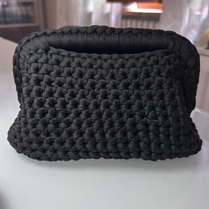 Può includere: Una pochette nera realizzata con un motivo a uncinetto denso e uniforme. La borsa presenta una struttura rettangolare con angoli arrotondati e un bordo intrecciato spesso lungo l'apertura superiore, creando un aspetto solido e strutturato.