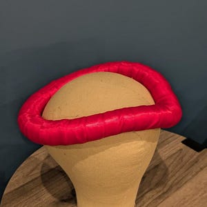 May include: A red satin headpiece, circular in shape, sits atop a tan mannequin head. The headpiece is a vibrant red, and the mannequin head is a neutral tan color. The background is a dark teal color.