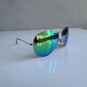 K&ouml;nnte beinhalten: Sonnenbrille im Aviator-Stil mit goldfarbenem Rahmen und irisierenden gr&uuml;n-blauen Gl&auml;sern. Die Gl&auml;ser reflektieren die Umgebung. Die Sonnenbrille liegt auf einer wei&szlig;en Oberfl&auml;che.