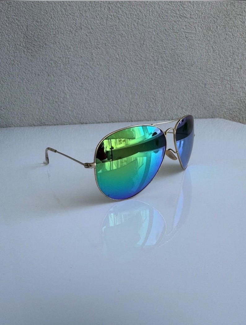 K&ouml;nnte beinhalten: Sonnenbrille im Aviator-Stil mit goldfarbenem Rahmen und irisierenden gr&uuml;n-blauen Gl&auml;sern. Die Gl&auml;ser reflektieren die Umgebung. Die Sonnenbrille liegt auf einer wei&szlig;en Oberfl&auml;che.