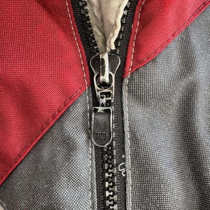 以下が含まれることがあります： ジャケットのジッパーのクローズアップ。ジッパーは黒で、シルバーのプルタブが付いています。ジャケットの生地は赤、グレー、ベージュです。ステッチは白です。