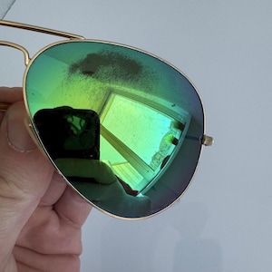 K&ouml;nnte beinhalten: Goldfarbene Aviator-Sonnenbrille mit gr&uuml;n get&ouml;nten Gl&auml;sern. Die Gl&auml;ser spiegeln eine helle, farbenfrohe Szene wider. Die Sonnenbrille hat einen Doppelsteg und ein klassisches Design.