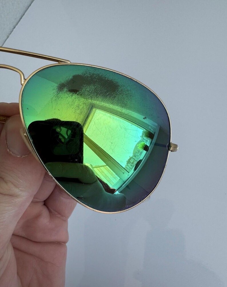 K&ouml;nnte beinhalten: Goldfarbene Aviator-Sonnenbrille mit gr&uuml;n get&ouml;nten Gl&auml;sern. Die Gl&auml;ser spiegeln eine helle, farbenfrohe Szene wider. Die Sonnenbrille hat einen Doppelsteg und ein klassisches Design.