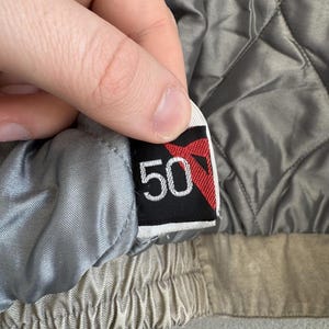 以下が含まれることがあります： 黒い背景に赤いデザインと白い数字の50が描かれたタグのクローズアップ。タグは、キルティング加工されたグレーのジャケットの裏地に取り付けられています。ジャケットの裏地は明るいグレーです。