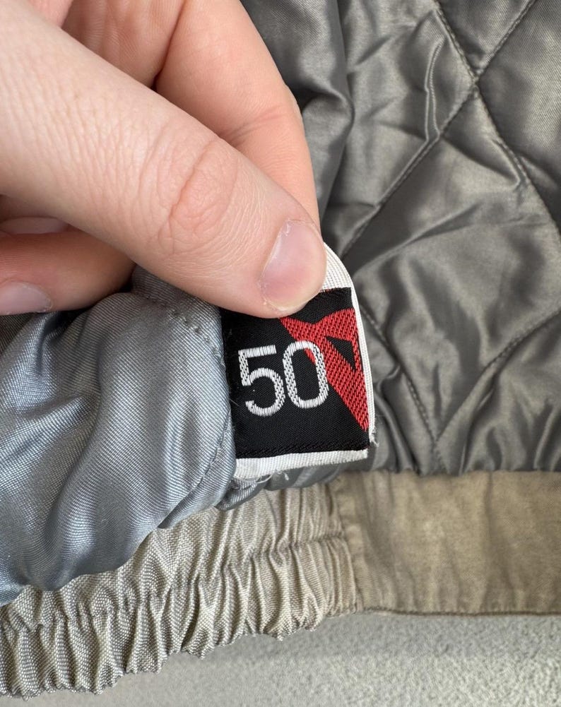 以下が含まれることがあります： 黒い背景に赤いデザインと白い数字の50が描かれたタグのクローズアップ。タグは、キルティング加工されたグレーのジャケットの裏地に取り付けられています。ジャケットの裏地は明るいグレーです。