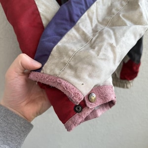 以下が含まれることがあります： ヴィンテージジャケットの袖のクローズアップ。袖は赤、白、紫の生地のパネルが特徴です。袖口は赤でピンクのトリムと2つの金色のボタンが付いています。