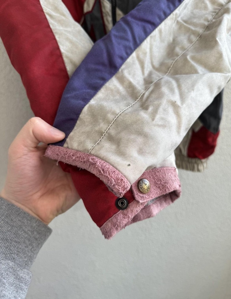 以下が含まれることがあります： ヴィンテージジャケットの袖のクローズアップ。袖は赤、白、紫の生地のパネルが特徴です。袖口は赤でピンクのトリムと2つの金色のボタンが付いています。