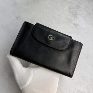 Może przedstawiać: Czarny skórzany portfel z klapką. Portfel posiada srebrny emblemat na klapie. Prostokątny portfel wykonany jest z gładkiej skóry. Portfel trzymany jest w rękawicy.