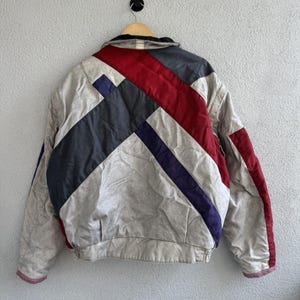 以下が含まれることがあります： 赤、ネイビー、グレーの幾何学模様が施された、ニュートラルカラーのヴィンテージジャケット。木製のハンガーにかけられ、明るいグレーの壁を背景にしています。ジャケットはリブ編みの裾と袖口が付いています。