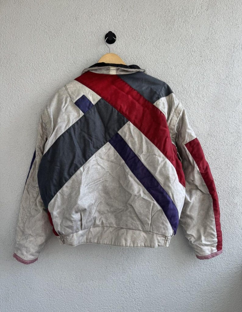 以下が含まれることがあります： 赤、ネイビー、グレーの幾何学模様が施された、ニュートラルカラーのヴィンテージジャケット。木製のハンガーにかけられ、明るいグレーの壁を背景にしています。ジャケットはリブ編みの裾と袖口が付いています。