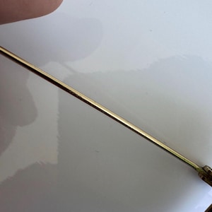 K&ouml;nnte beinhalten: Nahaufnahme eines goldfarbenen Metallb&uuml;gels einer Brille. Der B&uuml;gel ist d&uuml;nn und gerade, mit einer leichten Kr&uuml;mmung am Ende. Das Gestell liegt auf einer wei&szlig;en Oberfl&auml;che.