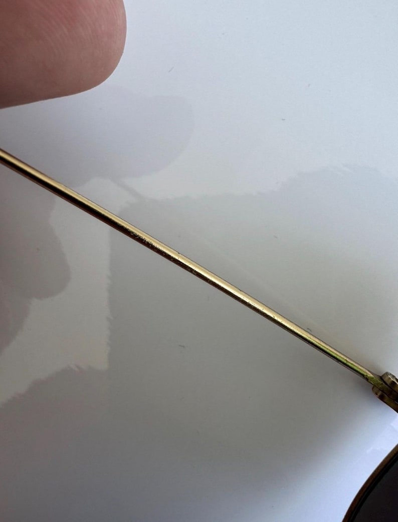 K&ouml;nnte beinhalten: Nahaufnahme eines goldfarbenen Metallb&uuml;gels einer Brille. Der B&uuml;gel ist d&uuml;nn und gerade, mit einer leichten Kr&uuml;mmung am Ende. Das Gestell liegt auf einer wei&szlig;en Oberfl&auml;che.