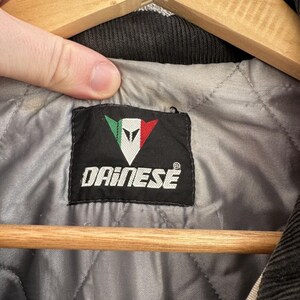 以下が含まれることがあります： 白字で「DAINESE」の文字と、緑と赤のアクセントが付いた白い三角形のロゴが入った黒いラベルのクローズアップ。 ラベルは、黒いコーデュロイの襟が付いた、ジャケットの灰色のキルティング裏地の中に縫い付けられています。