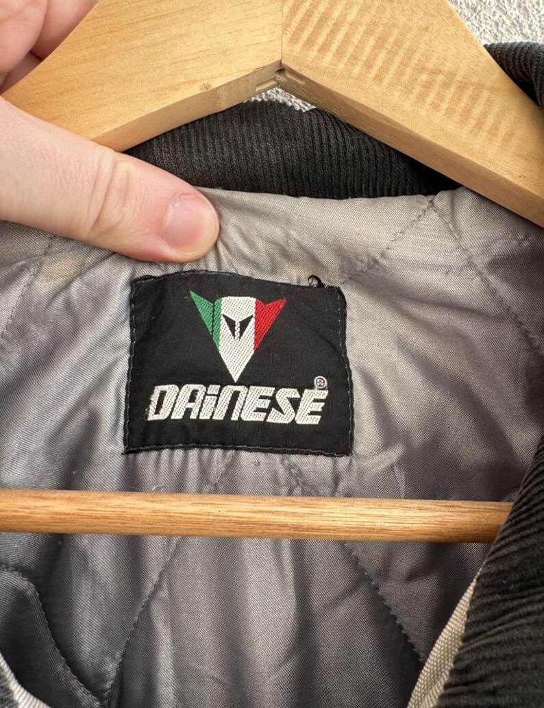 以下が含まれることがあります： 白字で「DAINESE」の文字と、緑と赤のアクセントが付いた白い三角形のロゴが入った黒いラベルのクローズアップ。 ラベルは、黒いコーデュロイの襟が付いた、ジャケットの灰色のキルティング裏地の中に縫い付けられています。