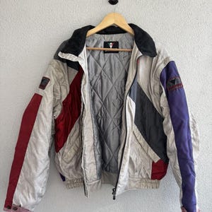 以下が含まれることがあります： 木製のハンガーに掛けられた、クリーム、赤、グレー、紫色のヴィンテージジャケット。ジャケットはキルティングの裏地、黒い襟、ジッパー開閉が特徴です。袖には赤と紫のアクセントがあります。