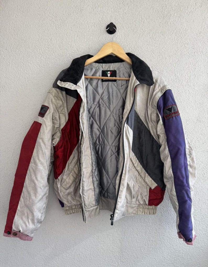 以下が含まれることがあります： 木製のハンガーに掛けられた、クリーム、赤、グレー、紫色のヴィンテージジャケット。ジャケットはキルティングの裏地、黒い襟、ジッパー開閉が特徴です。袖には赤と紫のアクセントがあります。