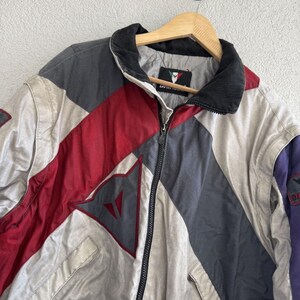 以下が含まれることがあります： グレー、赤、白のパネルを組み合わせた、カラーブロックデザインのヴィンテージジャケット。 黒い襟、フルレングスのジッパー、胸にロゴが付いています。 木製のハンガーに吊るされています。