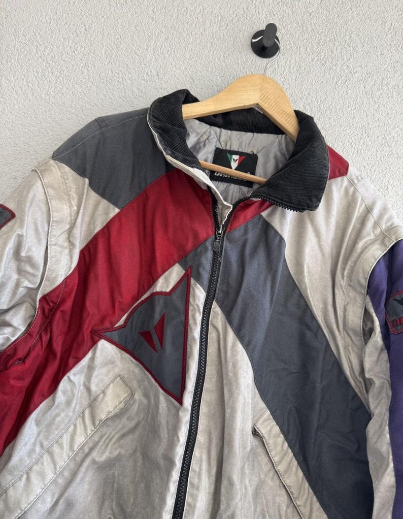 以下が含まれることがあります： グレー、赤、白のパネルを組み合わせた、カラーブロックデザインのヴィンテージジャケット。 黒い襟、フルレングスのジッパー、胸にロゴが付いています。 木製のハンガーに吊るされています。