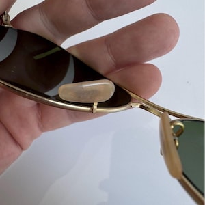 K&ouml;nnte beinhalten: Nahaufnahme einer Ray-Ban Sonnenbrille mit goldenem Metallrahmen und gr&uuml;nen Gl&auml;sern. Die Sonnenbrille hat ein klassisches Design mit Nasenpads und B&uuml;geln. Die Gl&auml;ser sind dunkel und bieten Schutz vor der Sonne.