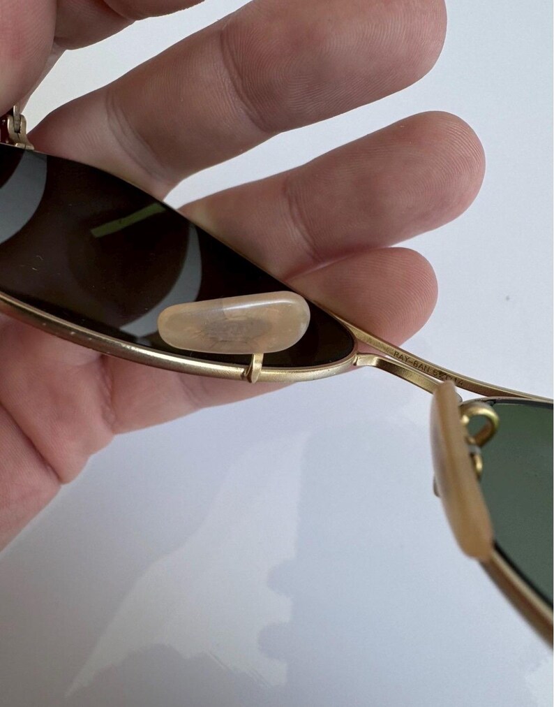 K&ouml;nnte beinhalten: Nahaufnahme einer Ray-Ban Sonnenbrille mit goldenem Metallrahmen und gr&uuml;nen Gl&auml;sern. Die Sonnenbrille hat ein klassisches Design mit Nasenpads und B&uuml;geln. Die Gl&auml;ser sind dunkel und bieten Schutz vor der Sonne.