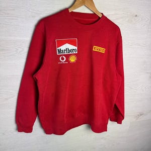 Könnte beinhalten: Rotes Sweatshirt mit dem Marlboro-Logo, Vodafone- und Shell-Logos auf der Vorderseite. Das Sweatshirt hat auch ein Pirelli-Logo. Das Kleidungsstück hängt an einem Holzbügel.