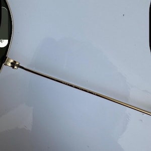 K&ouml;nnte beinhalten: Nahaufnahme einer Sonnenbrille mit goldfarbenem Metallrahmen und dunklen Gl&auml;sern. Der d&uuml;nne B&uuml;gel der Brille tr&auml;gt den Aufdruck "MADE IN ITALY". Die Brille ist vor einem wei&szlig;en Hintergrund platziert.