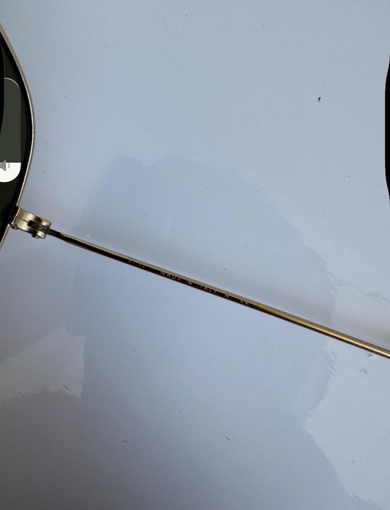 K&ouml;nnte beinhalten: Nahaufnahme einer Sonnenbrille mit goldfarbenem Metallrahmen und dunklen Gl&auml;sern. Der d&uuml;nne B&uuml;gel der Brille tr&auml;gt den Aufdruck "MADE IN ITALY". Die Brille ist vor einem wei&szlig;en Hintergrund platziert.