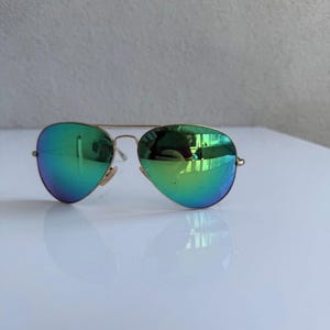 K&ouml;nnte beinhalten: Sonnenbrille im Aviator-Stil mit goldfarbenem Rahmen und verspiegelten gr&uuml;nen Gl&auml;sern. Die Gl&auml;ser haben einen Farbverlauf von Gr&uuml;n nach Blau. Die Sonnenbrille liegt auf einem wei&szlig;en Hintergrund.