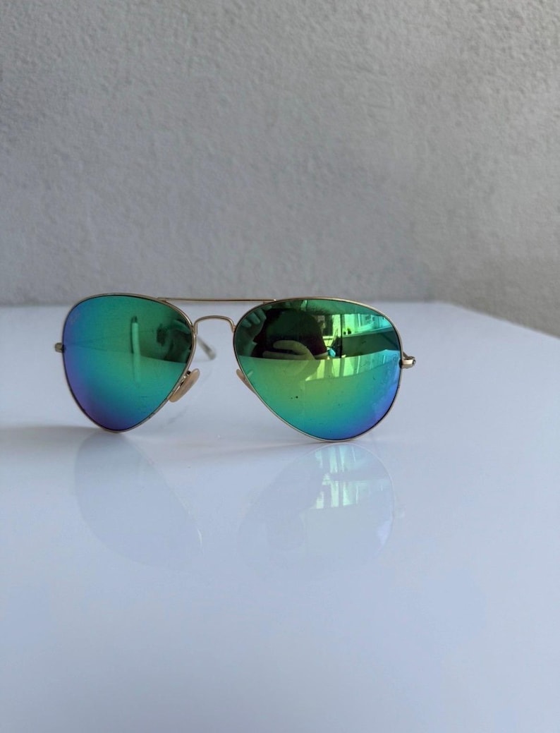 K&ouml;nnte beinhalten: Sonnenbrille im Aviator-Stil mit goldfarbenem Rahmen und verspiegelten gr&uuml;nen Gl&auml;sern. Die Gl&auml;ser haben einen Farbverlauf von Gr&uuml;n nach Blau. Die Sonnenbrille liegt auf einem wei&szlig;en Hintergrund.
