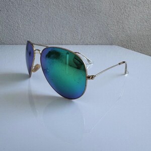 K&ouml;nnte beinhalten: Sonnenbrille im Aviator-Stil mit goldfarbenem Gestell und schillernden Gl&auml;sern, die von Blau zu Gr&uuml;n wechseln. Die Gl&auml;ser haben eine reflektierende Qualit&auml;t. Die Sonnenbrille liegt auf einer wei&szlig;en Oberfl&auml;che.