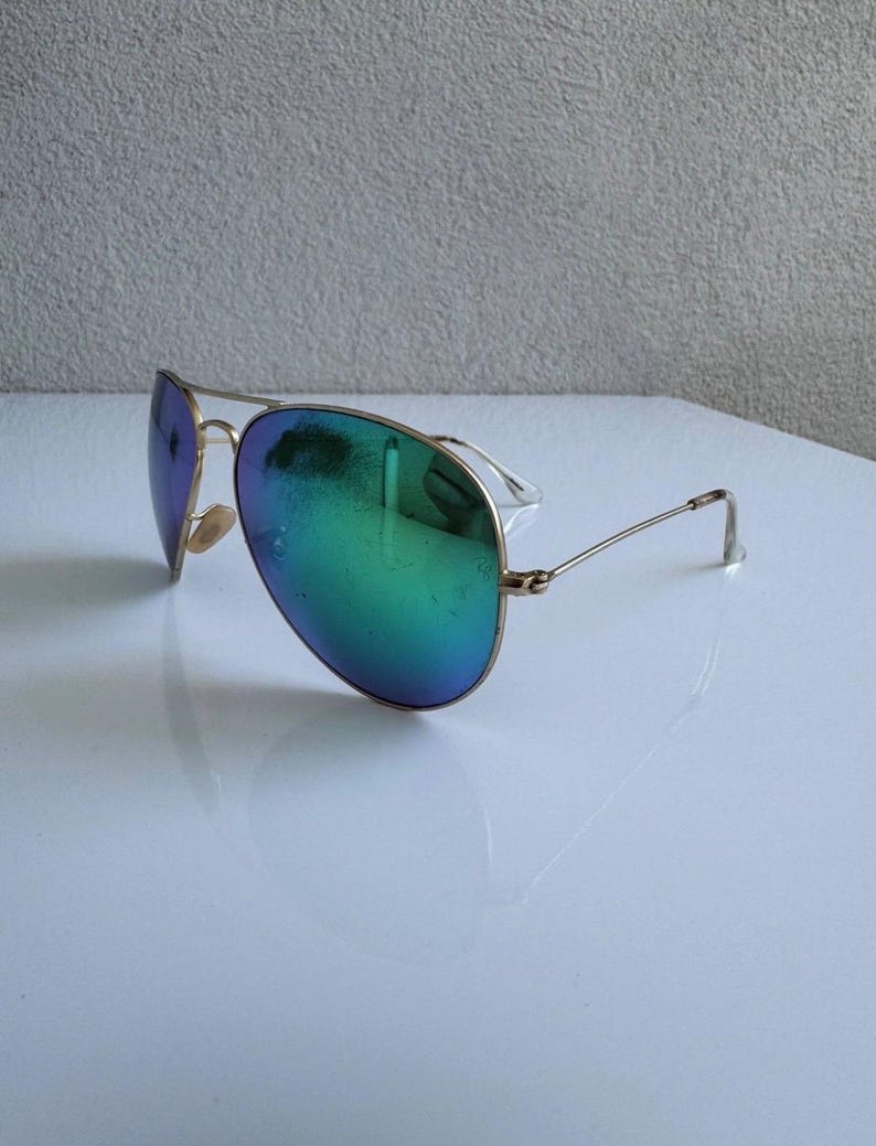 K&ouml;nnte beinhalten: Sonnenbrille im Aviator-Stil mit goldfarbenem Gestell und schillernden Gl&auml;sern, die von Blau zu Gr&uuml;n wechseln. Die Gl&auml;ser haben eine reflektierende Qualit&auml;t. Die Sonnenbrille liegt auf einer wei&szlig;en Oberfl&auml;che.