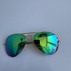 K&ouml;nnte beinhalten: Aviator-Sonnenbrille mit goldfarbenem Gestell und gr&uuml;n verspiegelten Gl&auml;sern. Die Gl&auml;ser spiegeln die Umgebung wider. Die Sonnenbrille hat eine klassische Tropfenform und einen Doppelsteg.