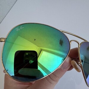K&ouml;nnte beinhalten: Goldfarbene Aviator-Sonnenbrille mit gr&uuml;n-blauen Farbverlaufsgl&auml;sern. Das Ray-Ban-Logo ist auf dem oberen linken Glas zu sehen. Die Gl&auml;ser spiegeln die Umgebung wider.