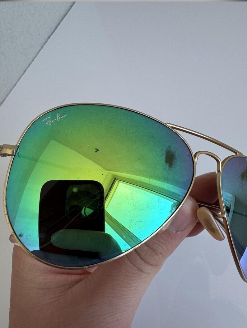 K&ouml;nnte beinhalten: Goldfarbene Aviator-Sonnenbrille mit gr&uuml;n-blauen Farbverlaufsgl&auml;sern. Das Ray-Ban-Logo ist auf dem oberen linken Glas zu sehen. Die Gl&auml;ser spiegeln die Umgebung wider.