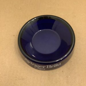 Könnte beinhalten: Ein runder, tiefblauer Glasaschenbecher mit dunklem Rand. Der Aschenbecher trägt den weißen Schriftzug "Ayingger Bräu Lager". Der Aschenbecher liegt auf einer hellbraunen Oberfläche.