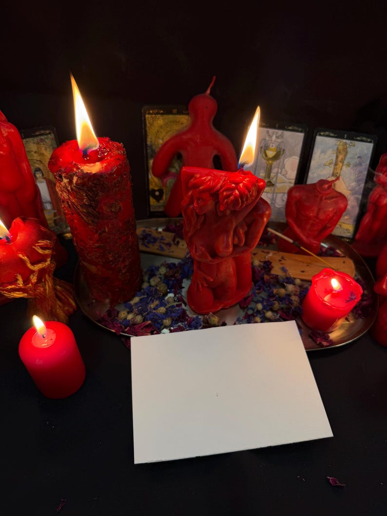 Puede incluir: Varias velas rojas de diferentes formas, algunas encendidas con llamas. Una tarjeta blanca en blanco en primer plano. Flores secas y cartas del tarot crean un ambiente m&iacute;stico.