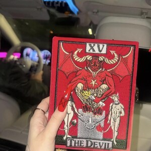 A person is holding a red and black tarot card with the text "THE DEVIL" on it, which is a major arcana card from the tarot deck.