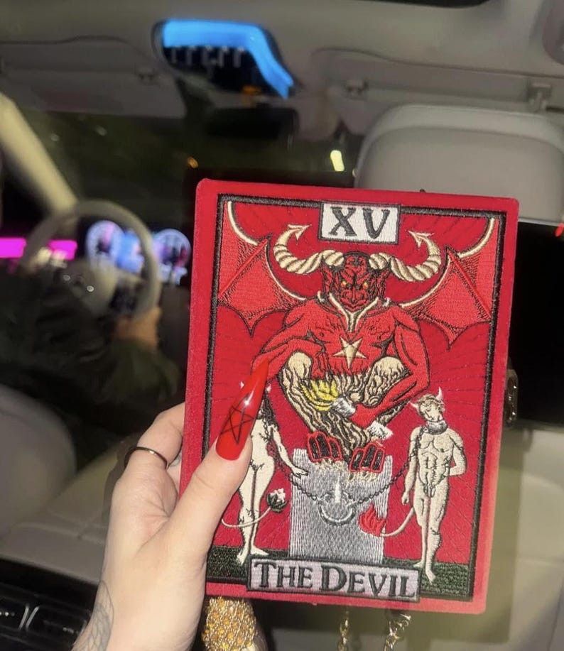 A person is holding a red and black tarot card with the text "THE DEVIL" on it, which is a major arcana card from the tarot deck.
