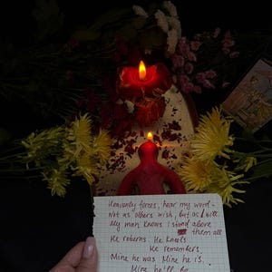 Puede incluir: Una vela roja encendida con forma de persona, con una nota manuscrita delante. La nota contiene texto. Flores amarillas y rosas son visibles en el fondo.