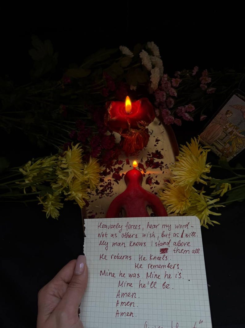 Puede incluir: Una vela roja encendida con forma de persona, con una nota manuscrita delante. La nota contiene texto. Flores amarillas y rosas son visibles en el fondo.
