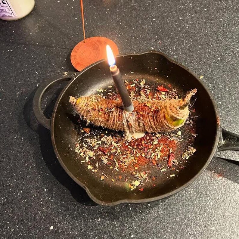 A black frying pan with a lit candle inside, surrounded by spices and herbs, on a dark surface.