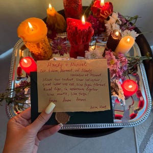 A hand holding a handwritten note with a warm, intimate message, surrounded by a tray of lit candles and flowers, creating a cozy and romantic atmosphere.