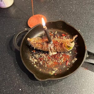 A black frying pan with a lit candle inside, surrounded by spices and herbs, on a dark surface.
