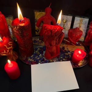 May include: A collection of red candles of various shapes and sizes, some lit with flames. A blank white card is in the foreground. Dried flowers and tarot cards are in the background, creating a mystical atmosphere.