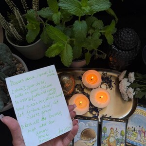 May include: A spiritual scene featuring lit candles, tarot cards, and fresh mint plants. A handwritten note with text is held in the foreground. The scene is set on a gold tray with a dark background.