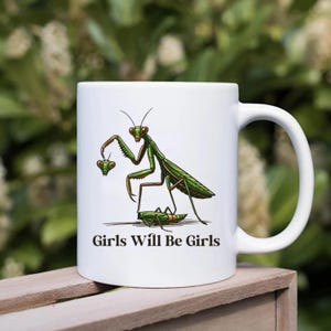 May include: White ceramic mug featuring a green and brown illustrated praying mantis holding two smaller mantises. The text "Girls Will Be Girls" is printed below the illustration. The mug has a curved handle.