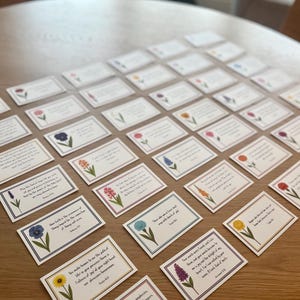 May include: A collection of rectangular cards with floral illustrations and text, arranged on a wooden surface. Each card features a different flower and a quote. The cards have a white background with a colored border.