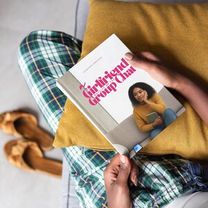 Puede incluir: Una persona sostiene un libro titulado "Girlfriend Group Chat" con una portada rosa y blanca. El libro descansa sobre un cojín amarillo. La persona lleva un pantalón de pijama a cuadros verdes y azules.
