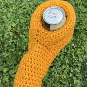 Puede incluir: Un portabotellas de ganchillo amarillo mostaza. El portabotellas est&aacute; dise&ntilde;ado para contener una lata de bebida de tama&ntilde;o est&aacute;ndar. El portabotellas est&aacute; hecho de un hilo grueso y las puntadas son visibles. La lata es plateada con una etiqueta blanca que dice "Sin cafe&iacute;na".