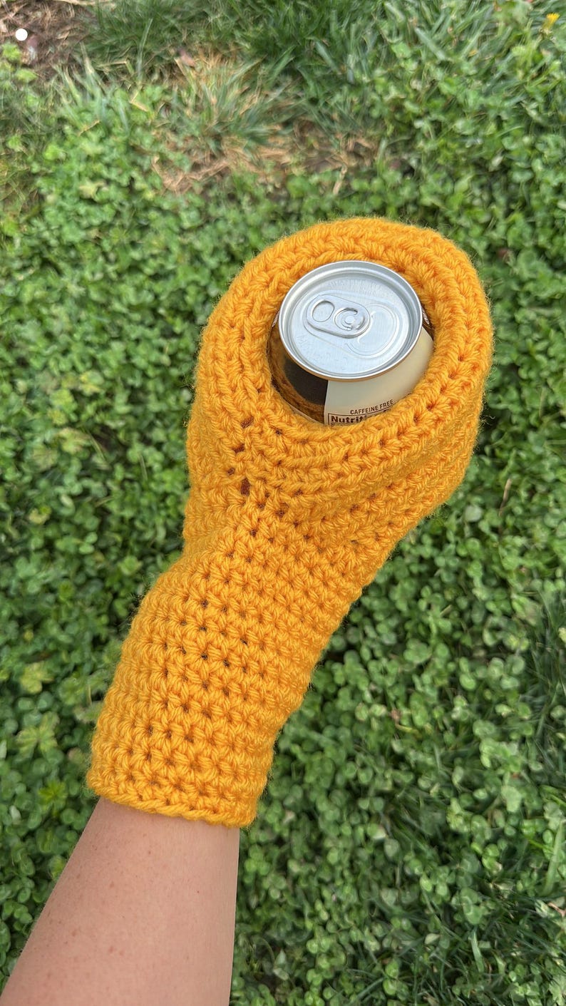 Puede incluir: Un portabotellas de ganchillo amarillo mostaza. El portabotellas est&aacute; dise&ntilde;ado para contener una lata de bebida de tama&ntilde;o est&aacute;ndar. El portabotellas est&aacute; hecho de un hilo grueso y las puntadas son visibles. La lata es plateada con una etiqueta blanca que dice "Sin cafe&iacute;na".